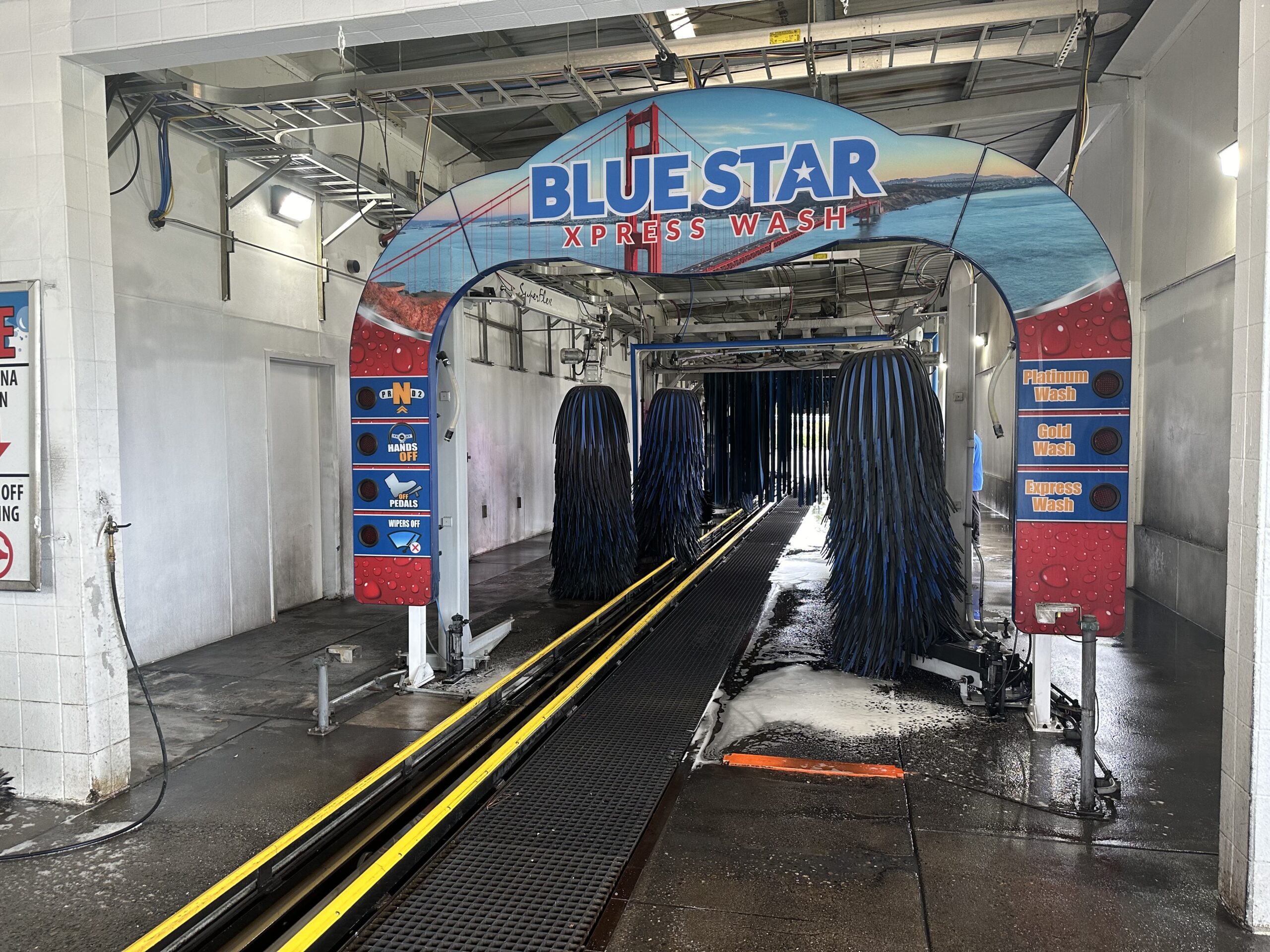 Inside a Blue Star Xpress Wash tunnel: three large blue mitter brushes hang over the conveyor track, a Golden Gate Bridge-themed entry arch labeled 'BLUE STAR XPRESS WASH' is visible, and Platinum, Gold, and Express wash signs line the right wall
